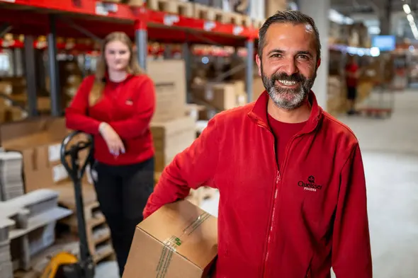 A smiling man in a red sweater holds a box in a warehouse, while a woman in a similar outfit stands behind him, leaning against a shelf. The background shows stacks of boxes and warehouse shelving.