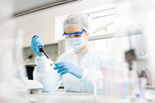 A person in a lab coat, gloves, goggles, and a mask is using a pipette to conduct an experiment in a laboratory setting. The background features lab equipment and glassware, emphasizing a scientific environment.
