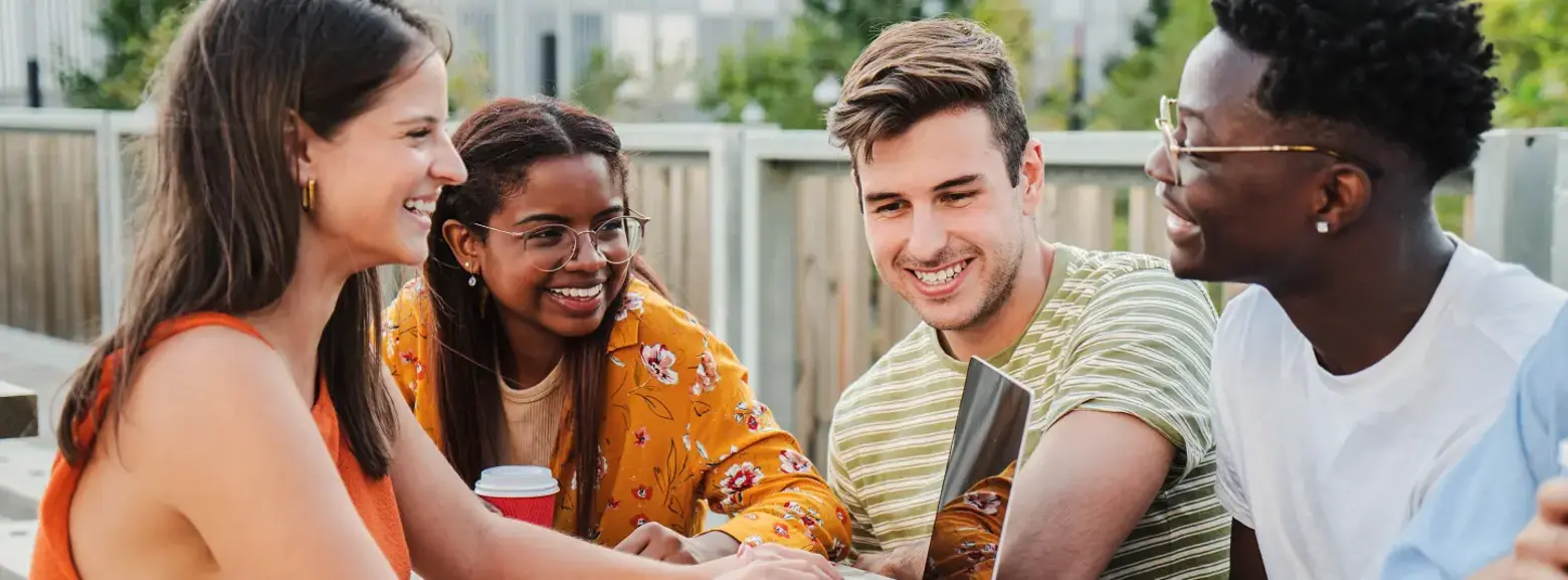 مجموعة من أربعة شبان يجلسون حول طاولة خشبية في الهواء الطلق، ينخرطون في نقاش حيوي وهم ينظرون إلى حاسوب محمول. يبدو أنهم يتعاونون أو يدرسون معًا، بابتسامات وأجواء من الرفاقية. أحدهم يحمل فنجان قهوة.