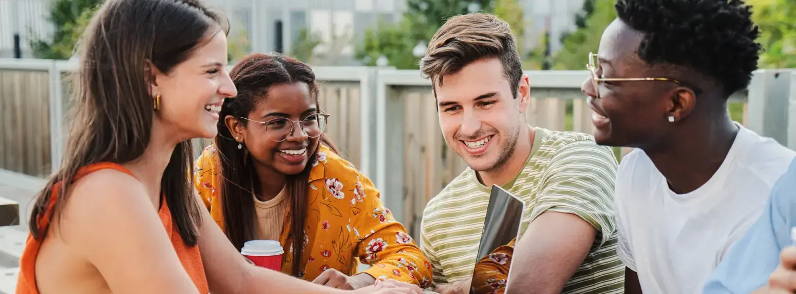 A group of four young adults sit around a wooden table outdoors, engaged in a lively discussion while looking at a laptop. They appear to be collaborating or studying together, with smiles and an atmosphere of camaraderie. One person holds a coffee cup.