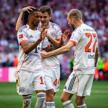 Three football players from Bayern Munich celebrate a goal. One player, wearing jersey number 14, is being congratulated by teammates, with one placing a hand on his head. They are wearing white and orange uniforms with the team logo visible. A crowd is blurred in the background.