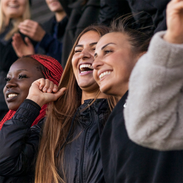 Three young women smile and cheer, expressing joy and excitement. They are dressed casually, with one wearing a black jacket and the others in light-colored tops. The background suggests a lively atmosphere, possibly at a sporting event or gathering.