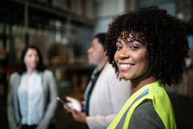A smiling woman with curly hair and a yellow safety vest stands in a warehouse setting, while two other women are engaged in conversation in the background. The atmosphere appears professional and collaborative.