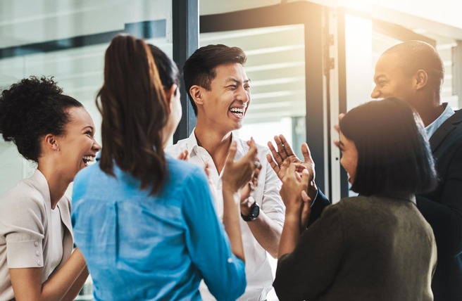 A diverse group of six people, including men and women, are laughing and celebrating in a bright, modern office setting. They are engaged in an animated conversation, clapping and smiling, exuding a sense of joy and camaraderie.