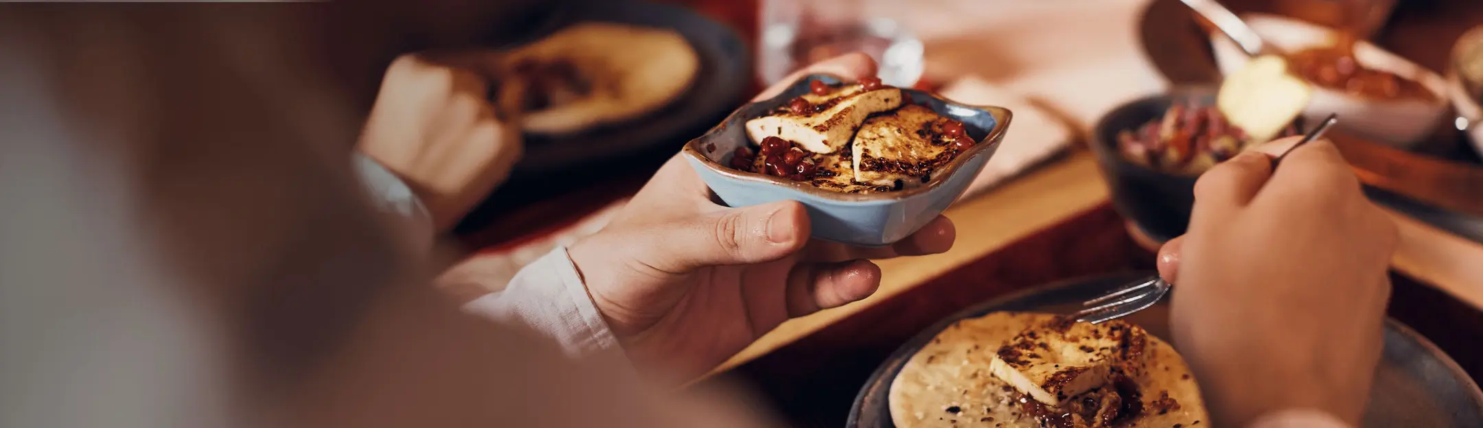 صورة تُظهر يد تُمسك بوعاء صغير يحتوي على قطع من الطعام المزينة بصلصة، بينما تُظهر اليد الأخرى ملعقة. في الخلفية، توجد أطباق طعام أخرى. الأجواء مريحة ومُريحة تتعلق بتناول الوجبة.