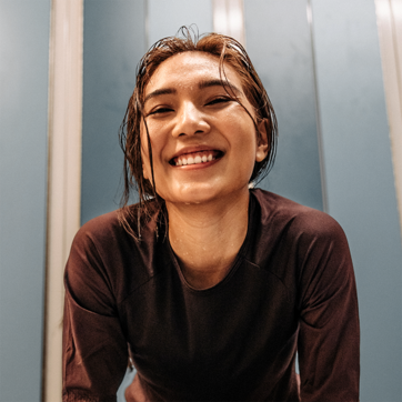 A smiling woman with wet hair leans forward, exuding joy and energy. She wears a dark shirt and stands against a backdrop of blue walls. Her expression conveys enthusiasm, possibly after engaging in physical activity.