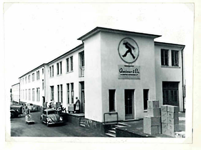 A black and white historic photograph of a building labeled "Quinivar & Co." featuring a logo of a person walking. The structure has multiple windows, a set of stairs, and boxes stacked nearby. Vintage cars are parked in front, with a few people visible outside.