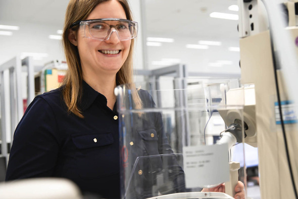 A smiling woman wearing safety glasses stands in a laboratory setting, next to a piece of lab equipment. She is dressed in a dark shirt and appears engaged in her work. The background shows shelves and equipment typical of a research environment.